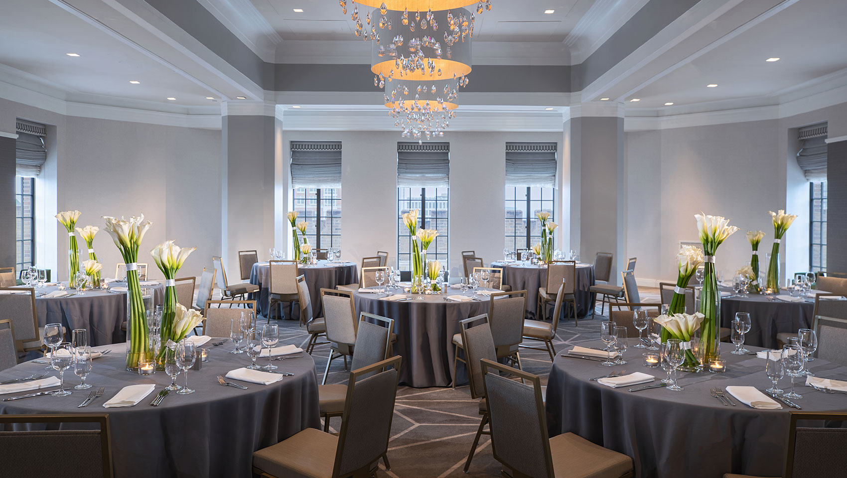 Kimpton Hotel Palomar Philadelphia ballroom set up for a social banquet with multiple round tables with floral arrangements placed in a sunlit room with surrounding windows overlooking views of the city