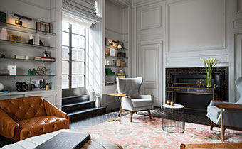 Kimpton Hotel Palomar Philadelphia Aia Library showing accent chairs surrounding a fireplace with bookshelves on background wall with large sunlit windows 