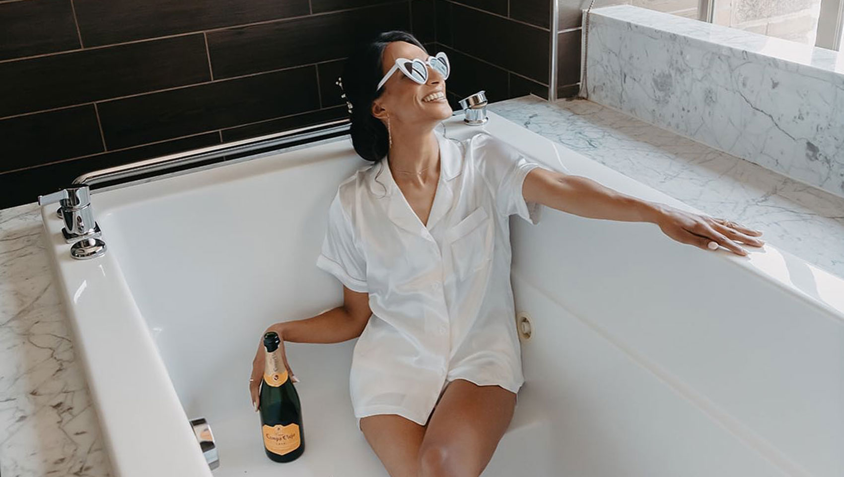 woman with sunglasses dressed in white, sitting in a spa bathtub and holding a bottle of champagne