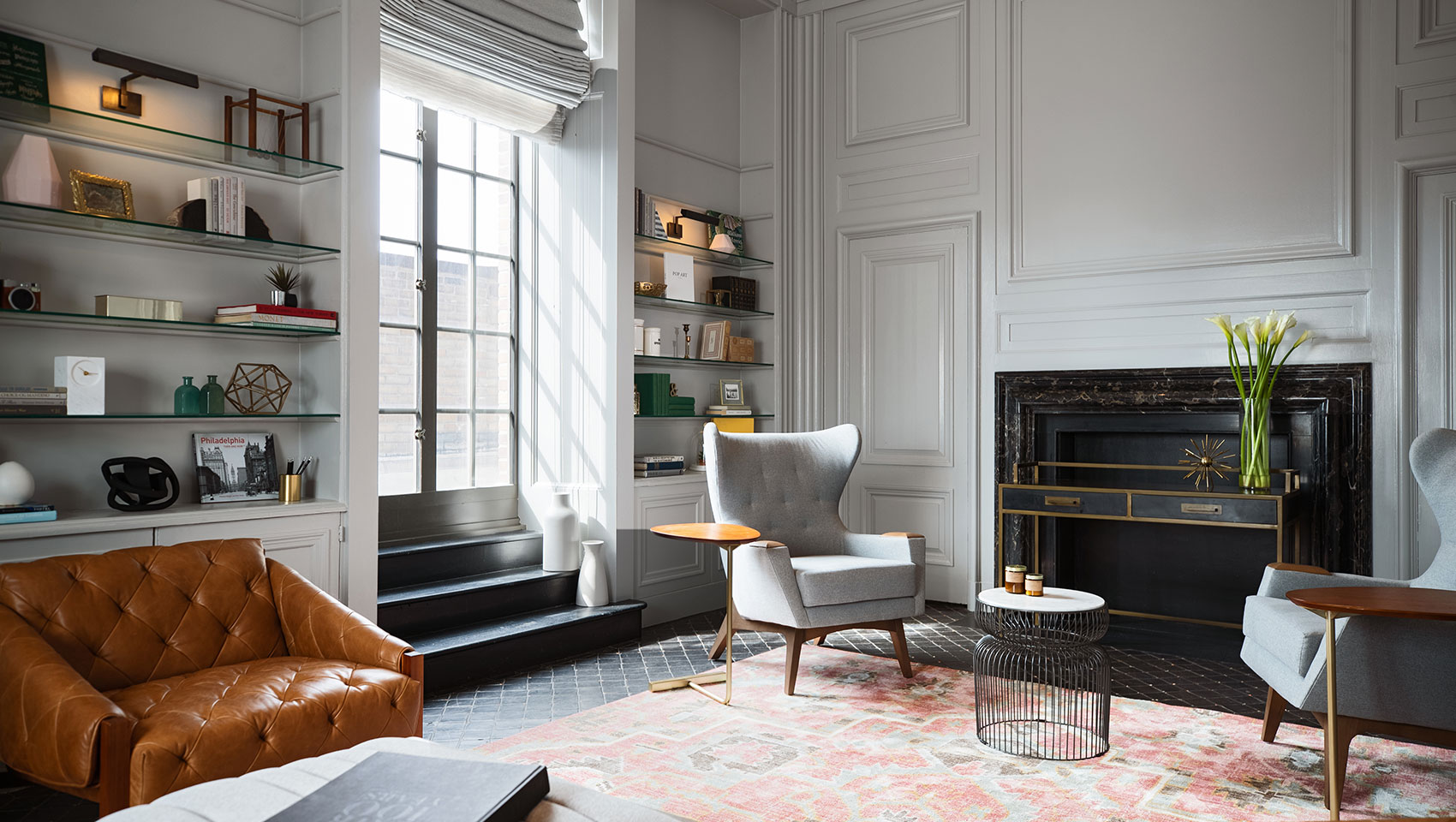 Kimpton Hotel Palomar Philadelphia Aia Library showing accent chairs surrounding a fireplace with bookshelves on background wall with large sunlit windows 