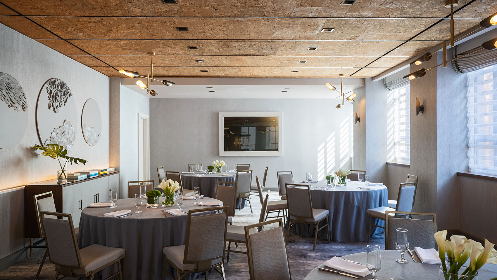 Social banquet setup in Tyng room with multiple round tables that have place settings and floral pieces in the center of the table, situated in a room with sunlit windows